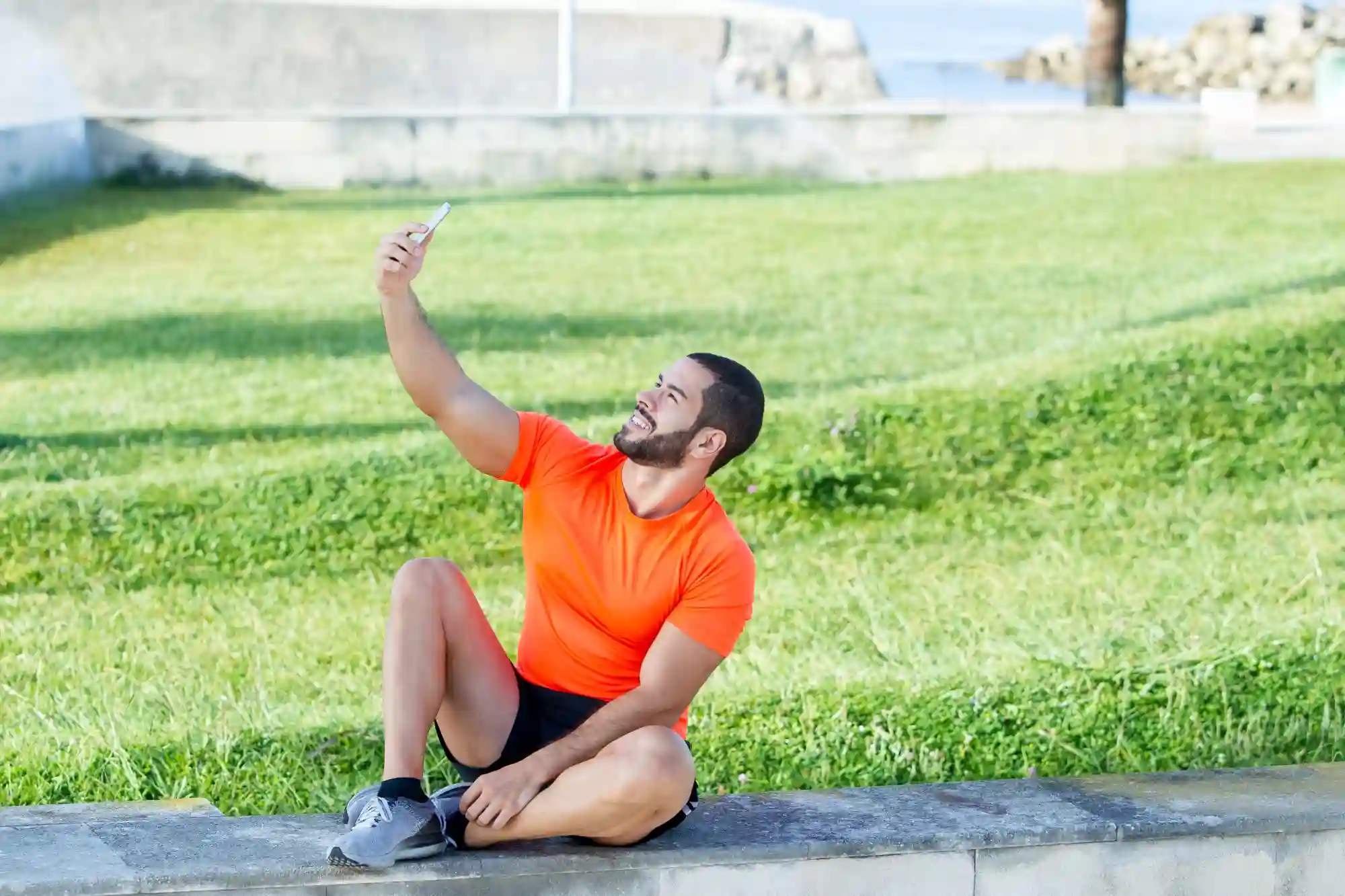 A man in athletic wear sitting on a stone ledge outdoors taking a selfie with his smartphone.