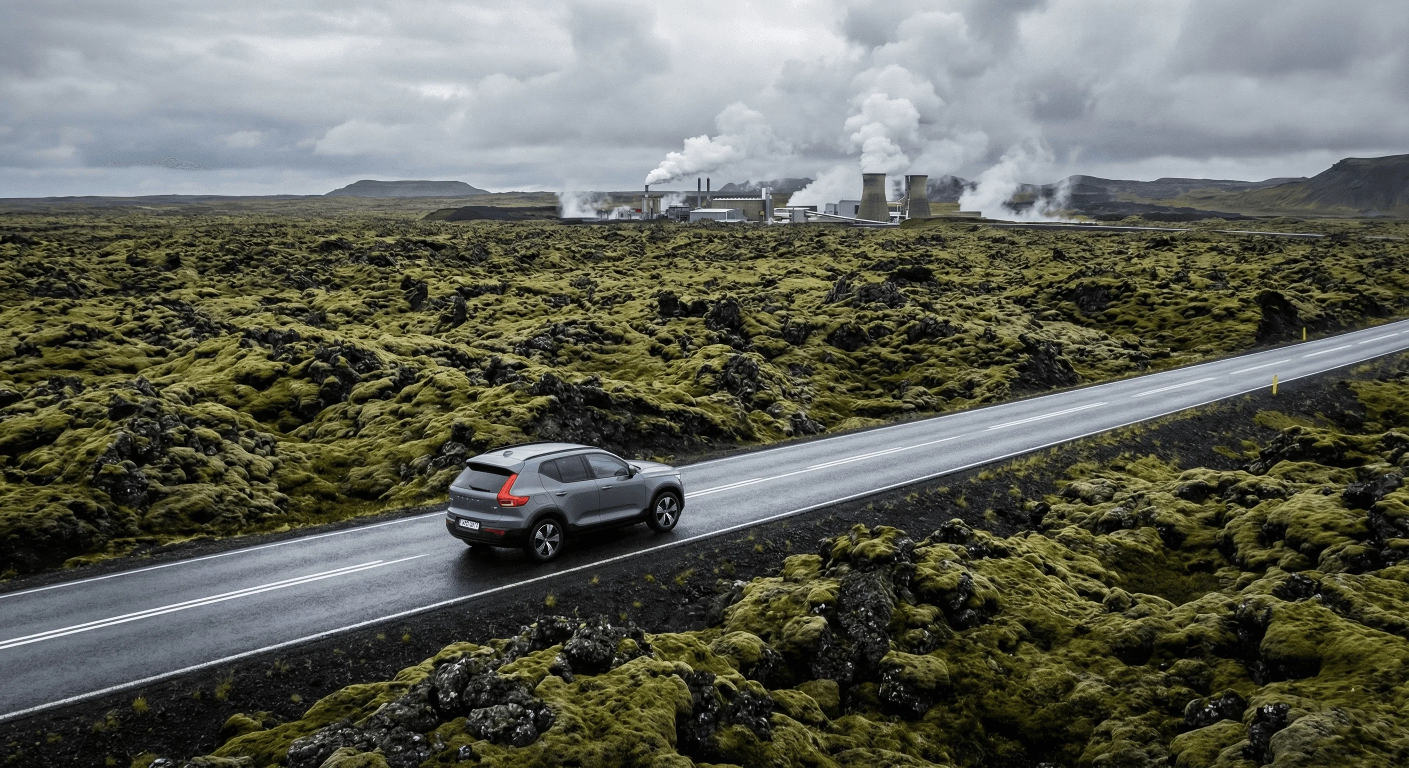 A silver SUV driving on a wet road winding through a green moss-covered lava field with geothermal steam in the background.