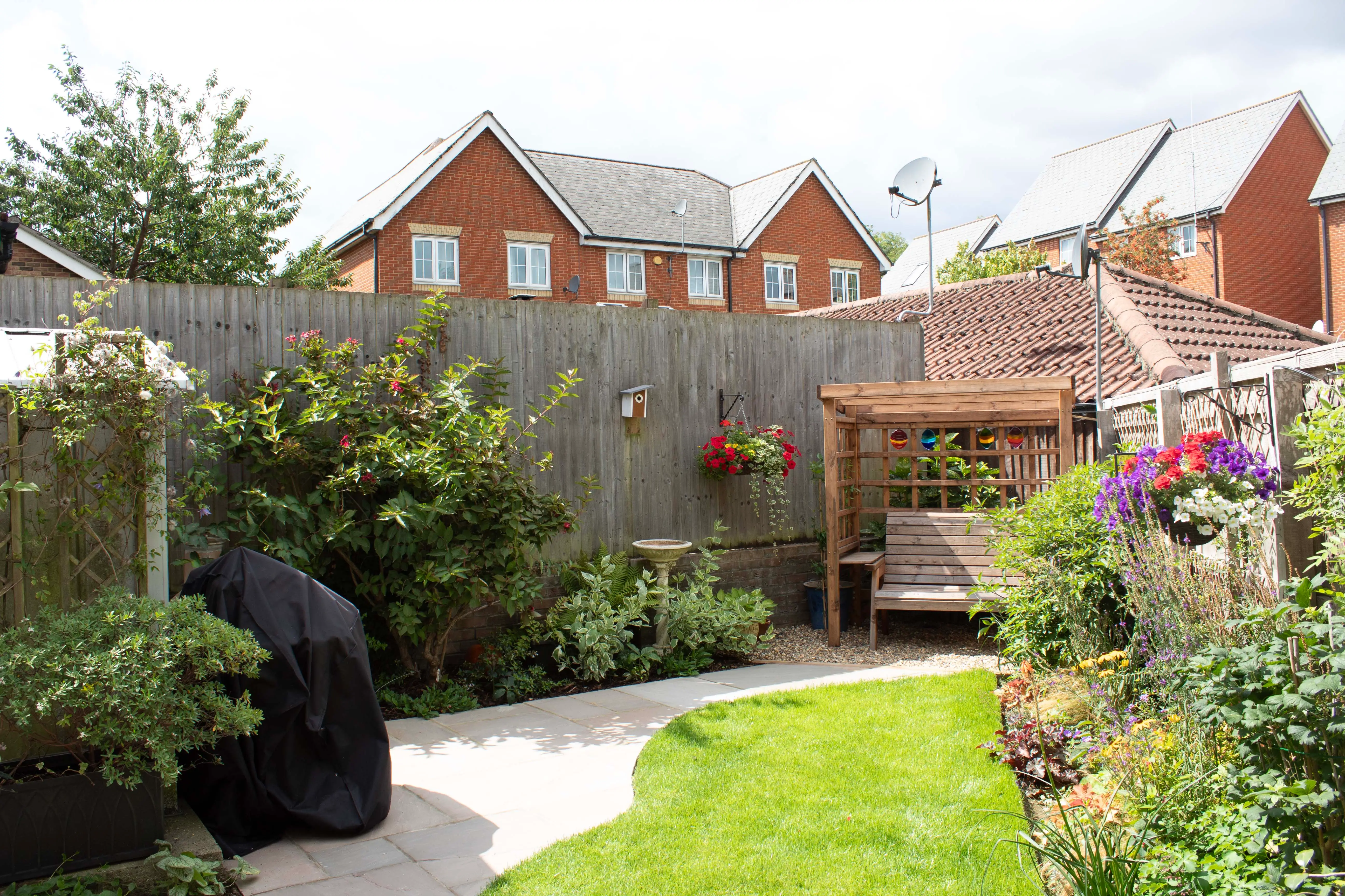 A sunny garden with green plants, a gravel path, a seating area, and houses in the background.