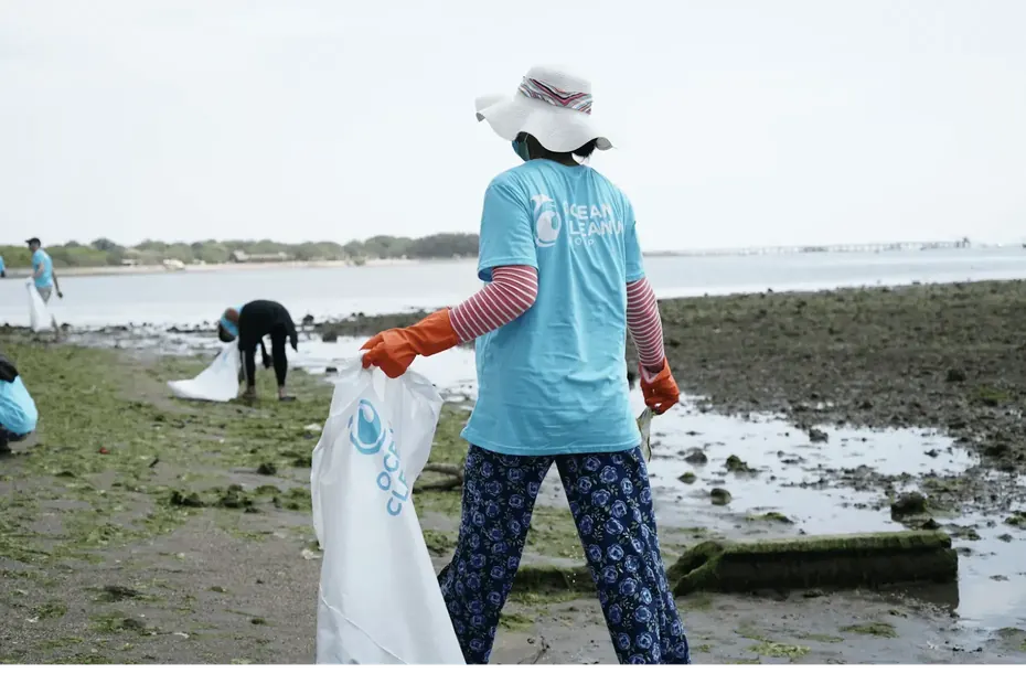 Volunteer in a sun hat collecting trash along a shoreline.