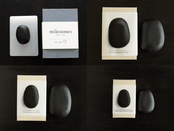 Four black stones rest on white and beige cards on a dark surface. The top left card displays a white rectangular object with a black stone. The top right card shows a black stone on a white card. The bottom left card has a black stone on a white card. The bottom right card displays two black stones on separate cards. Text on the cards reads “milestones,” “memory capsules,” and “MY COLLECTION FINDS AT A VENDOR.”