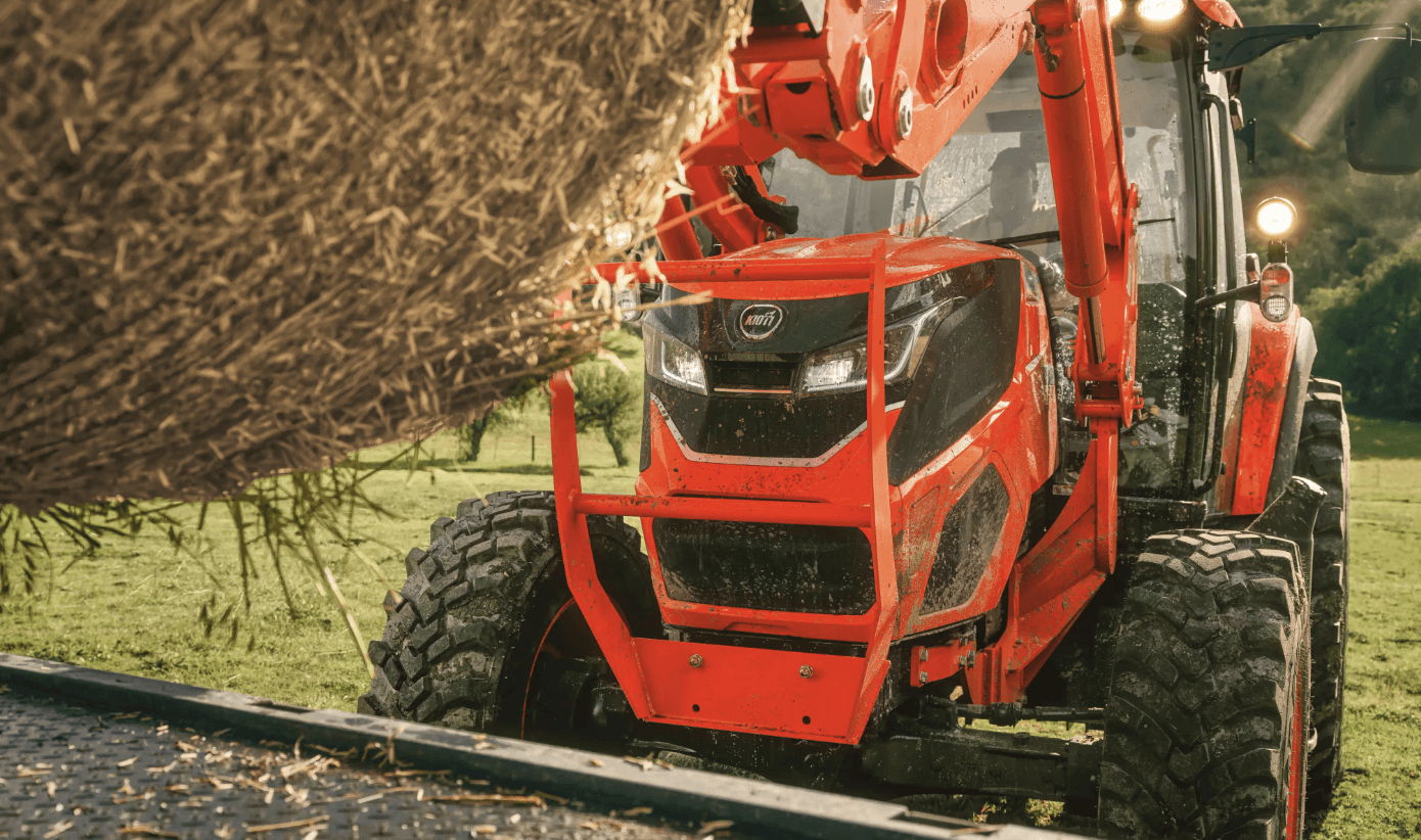 A KIOTI tractor loading a hay bale onto a flatbed truck.