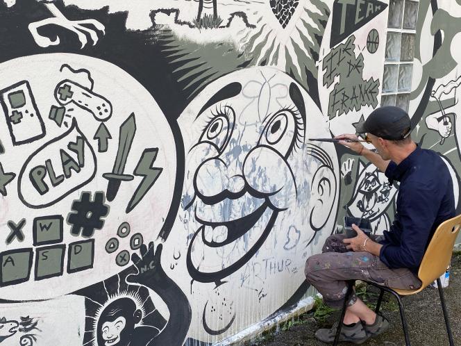 A man on a chair paints a grinning cartoon face among drawings of game controllers.