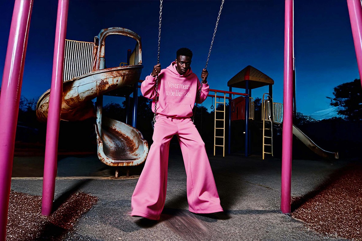 Model wearing pink oversized hoodie and pants standing by playground swing, Youth Distortion collection campaign.