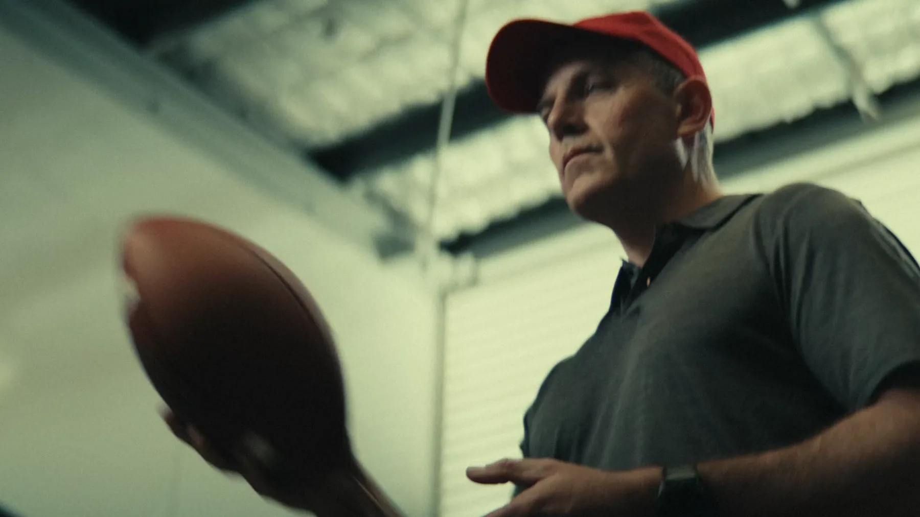 Low angle close-up of a coach wearing a red cap and gray polo shirt, tossing a football, standing under the exposed ceiling beams of an indoor facility.