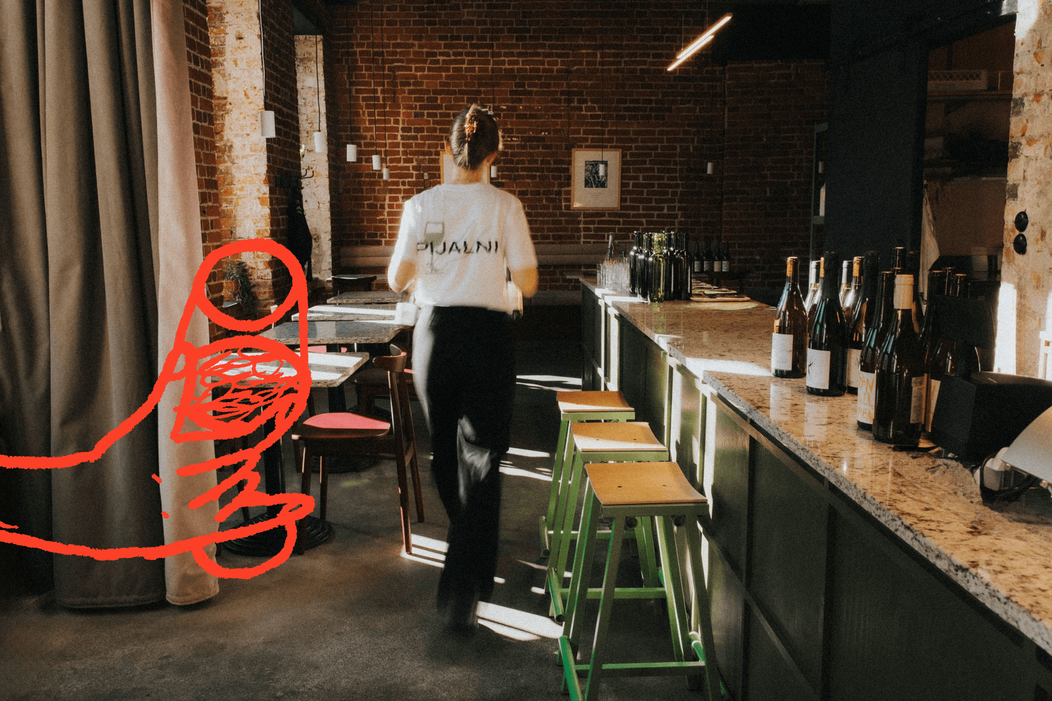 A person stands in a cozy wine bar with wooden decor, near colorful stools and a counter with coffee equipment.