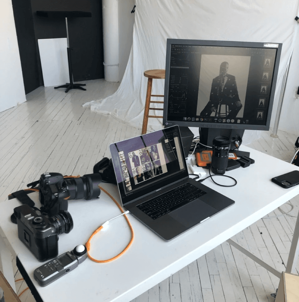 A white table with DSLR and medium-format cameras, laptop, and monitor displaying black-and-white portraits in a photo studio