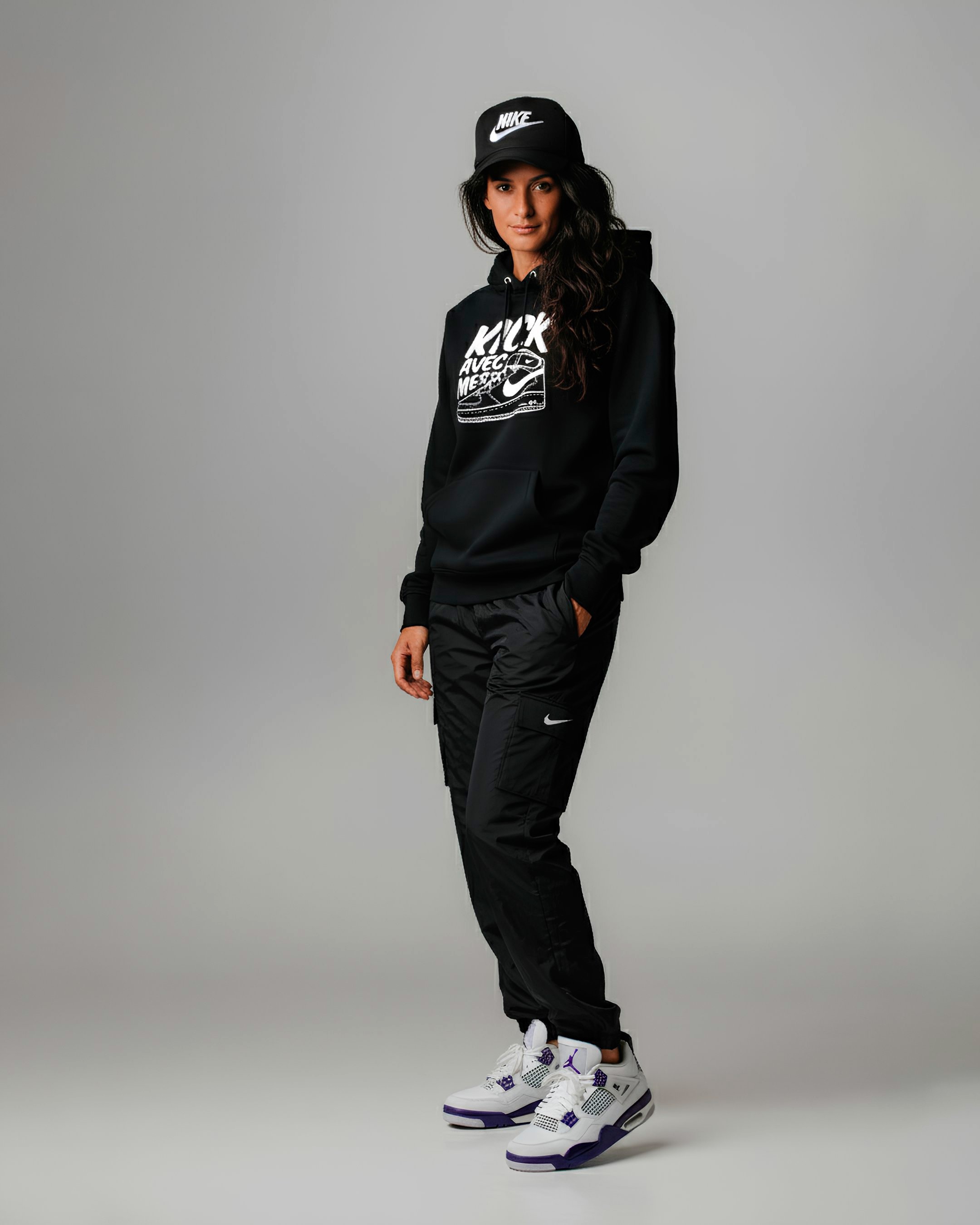Photo de studio en pleine longueur d'une jeune femme au style urbain et décontracté, portant une casquette noire 'Nike' et un sweat à capuche noir avec un grand motif blanc imprimé. Elle est habillée d'un pantalon cargo noir à fermeture éclair avec un petit logo Nike, et de sneakers blanches et violettes de type Jordan 4. Sa main gauche est dans la poche. L'arrière-plan est un fond uni gris clair.