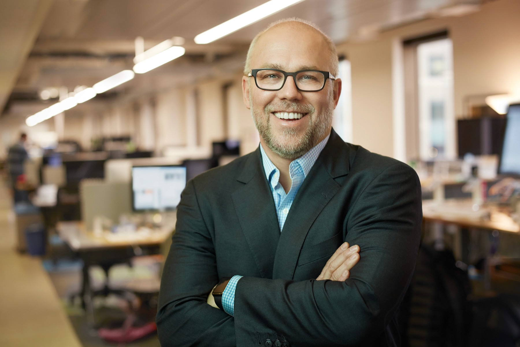 Confident headshot of bald man in glasses smiling inside open-plan tech office