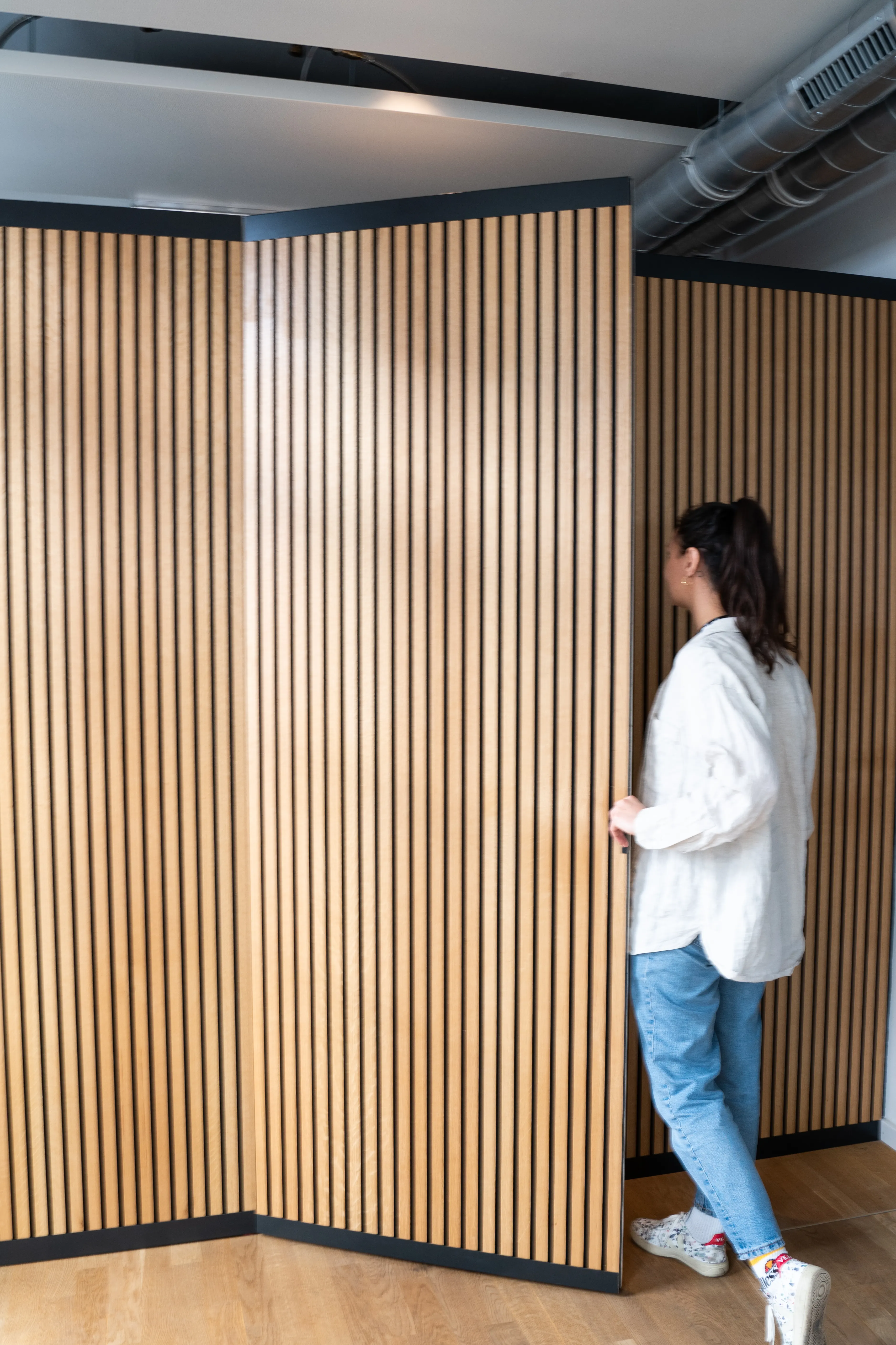 Person opening a folding partition made of oak acoustic slat panels with black trim at AI START-UP MUNICH, with exposed ventilation pipe at the ceiling.