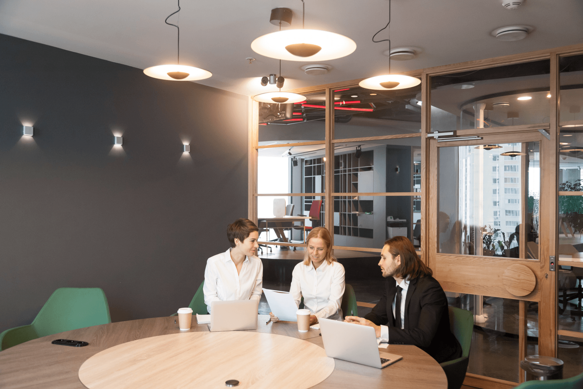 Three business professionals in a meeting room discussing documents at a round table.