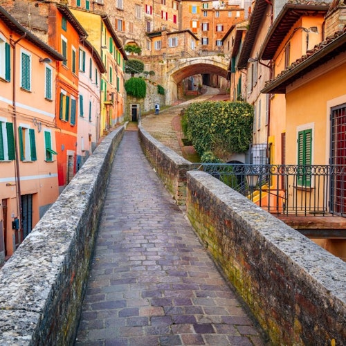 A stone-paved walkway leads through colorful buildings, with an arch and stairs in the background, under a bright sky.