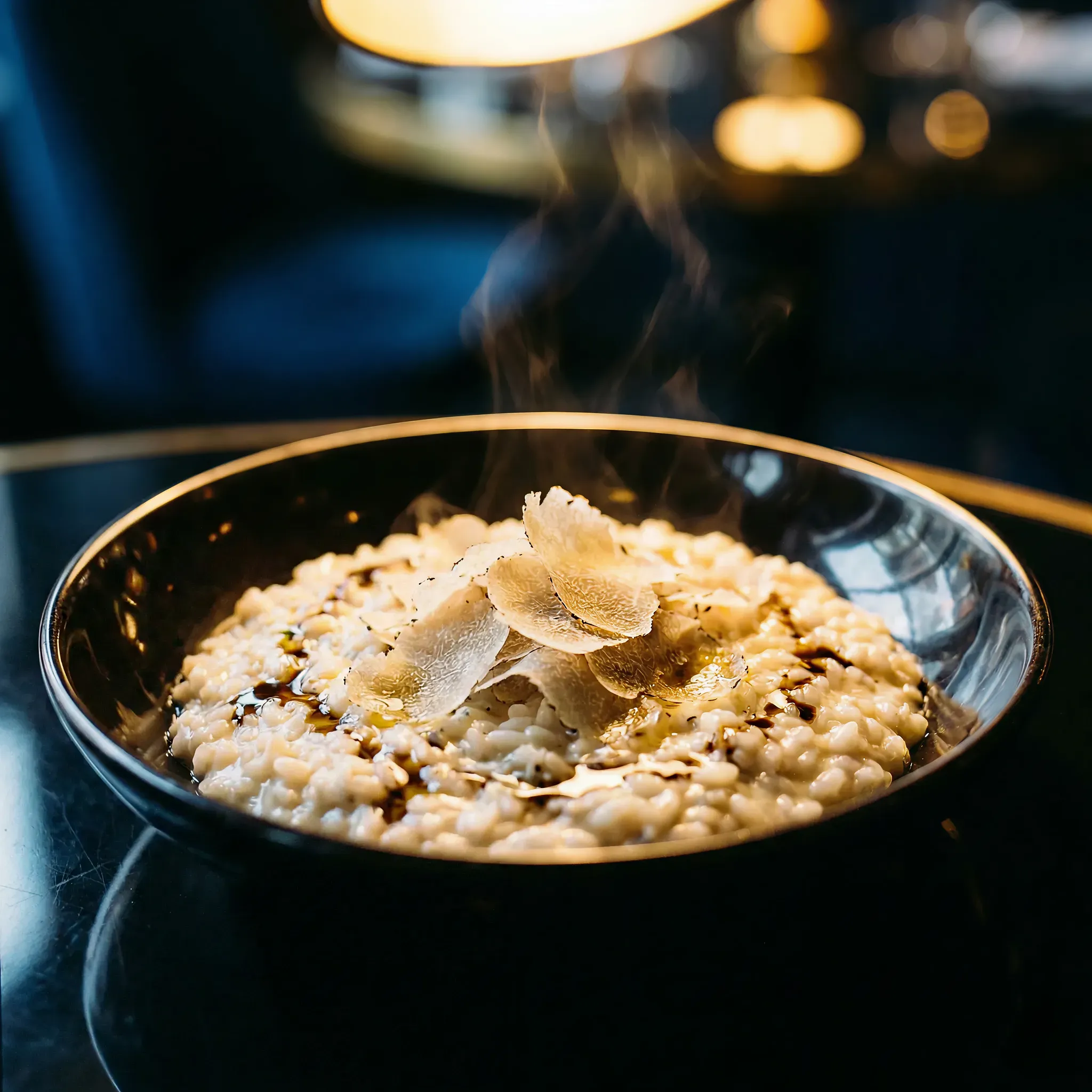 Steaming risotto topped with shaved truffles in a black bowl under warm lighting. The setting is elegant, evoking a refined dining experience.