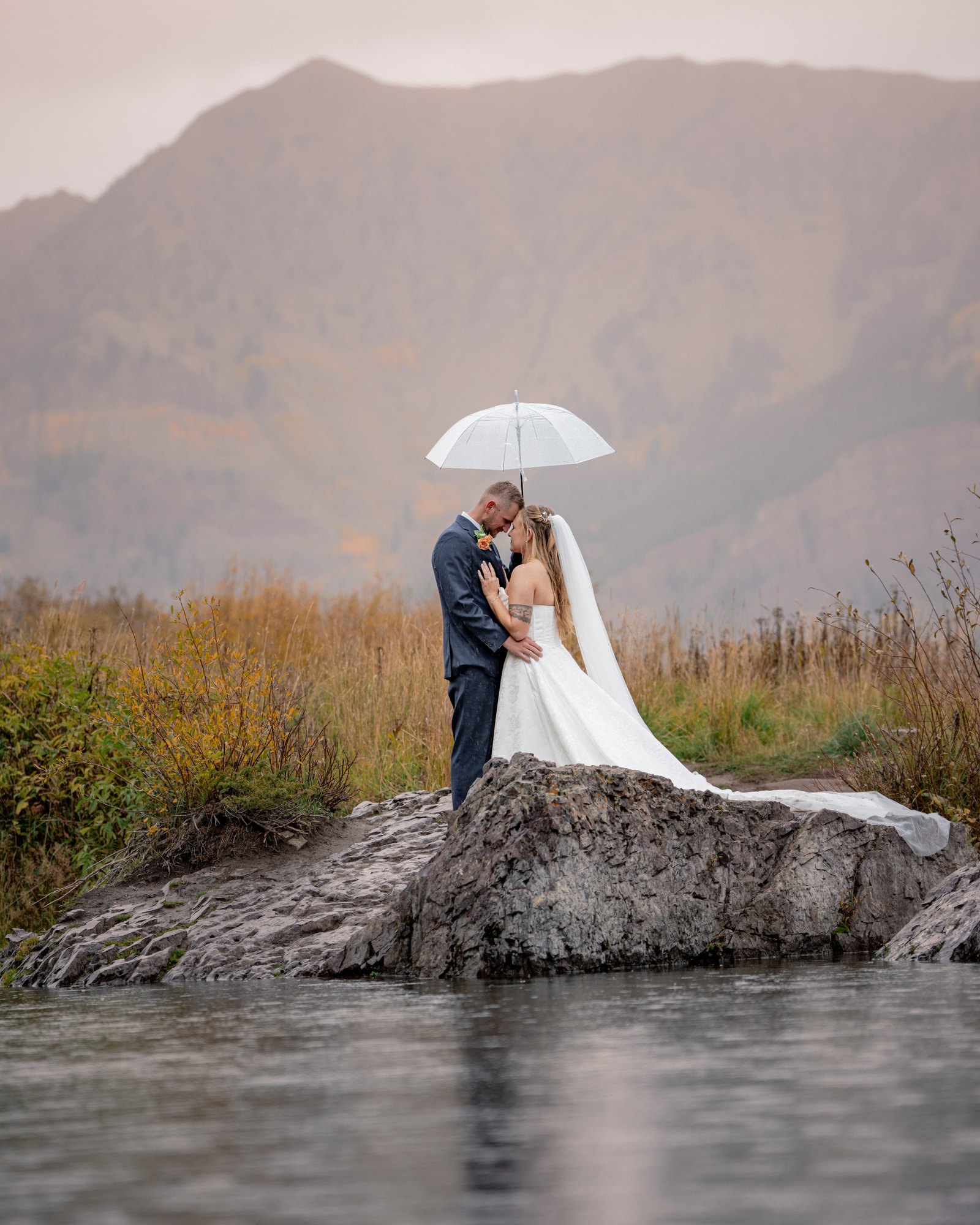 Wedding couple under a clear umbrella on a rock beside Maroon Lake