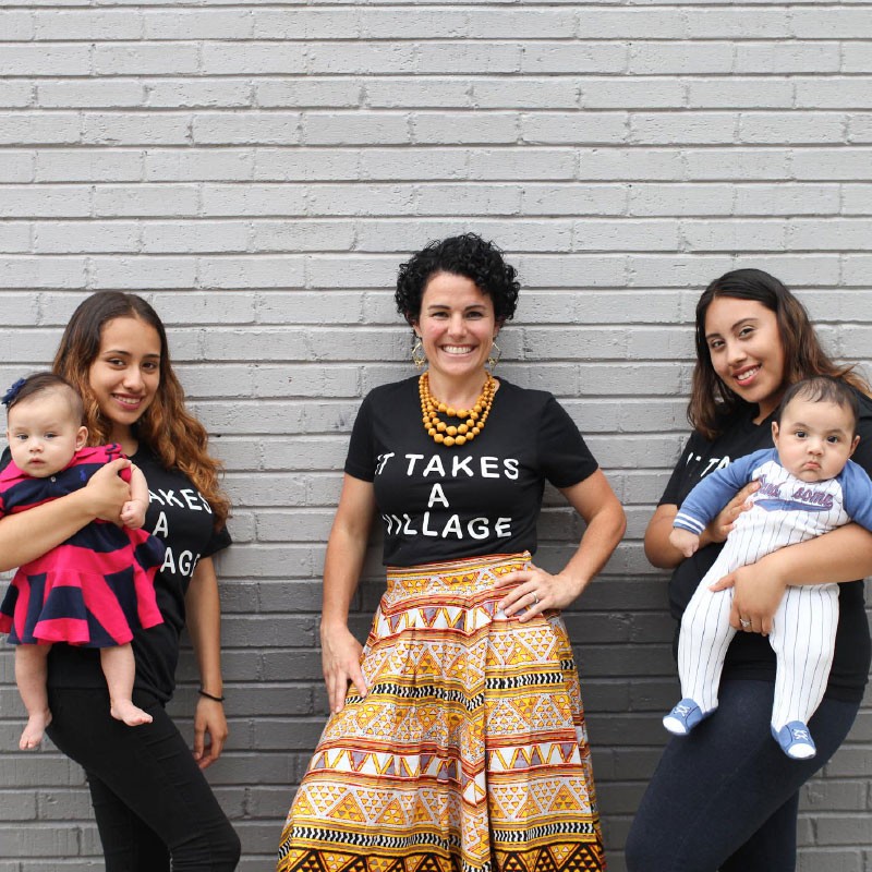 Four women, two holding babies, stand in front of a brick wall. They wear black t-shirts that say "It takes a village".