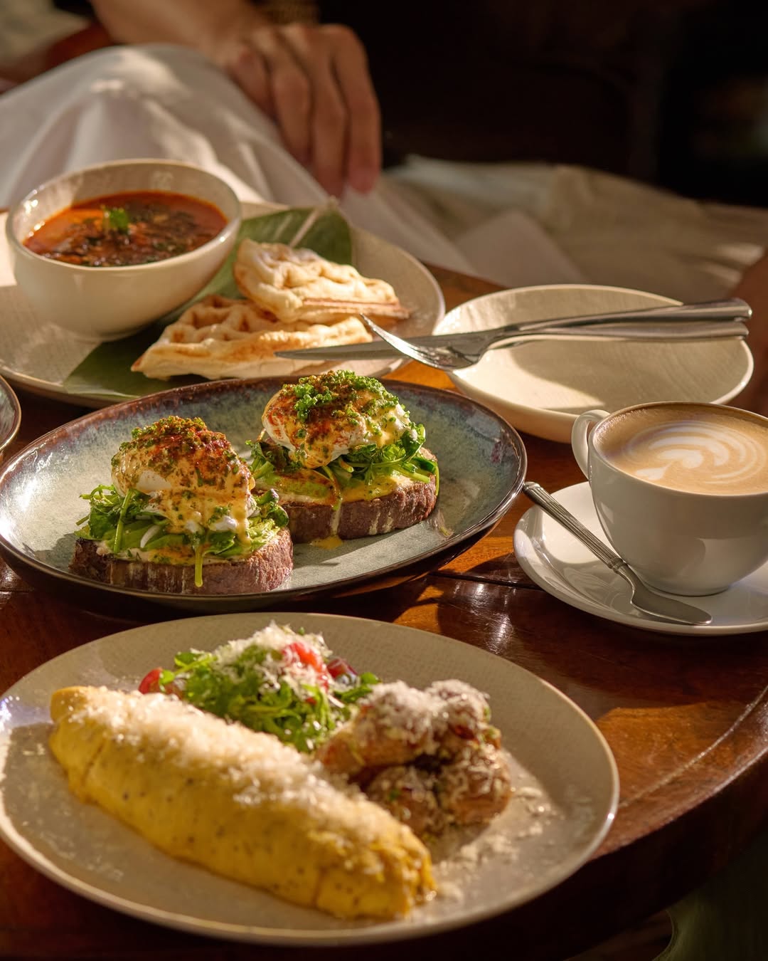 A beautifully arranged breakfast scene featuring two plates of gourmet food, including avocado toast with poached eggs, a fluffy omelette with salad, a bowl of soup, flatbreads, and a cup of latte, all placed on a wooden table.