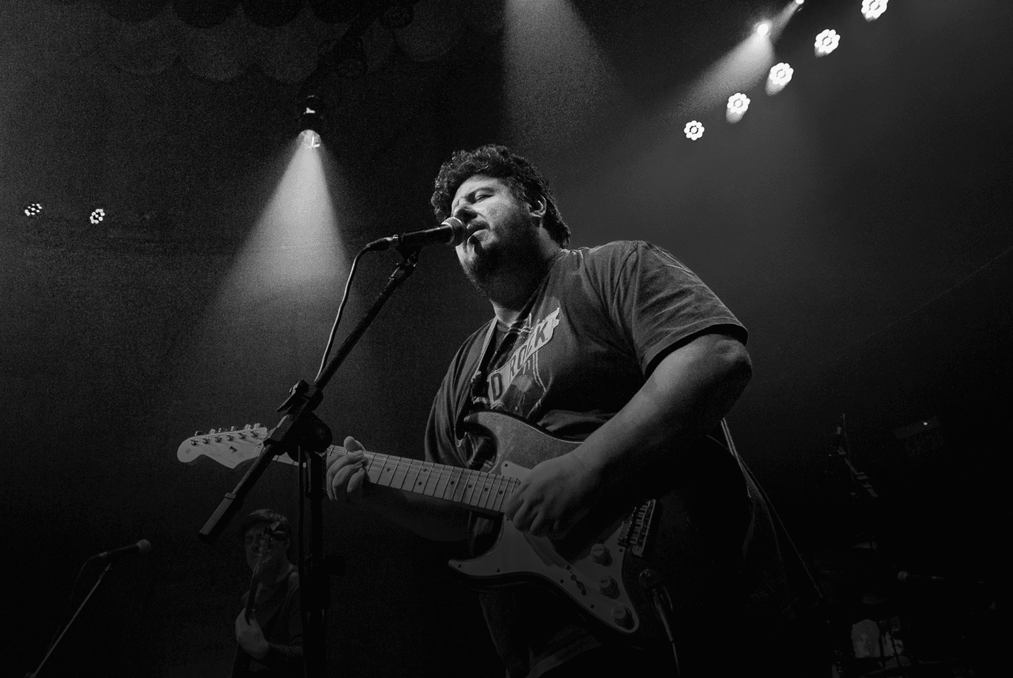 Guitarrista de blues internacional se apresentando ao vivo no palco, cantando ao microfone e tocando guitarra durante show.
