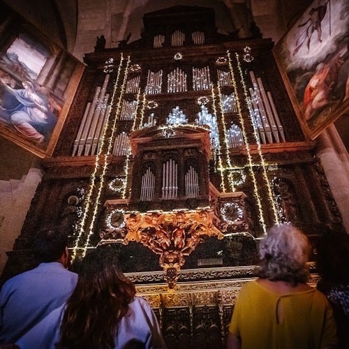 Four people stand facing an ornate church organ adorned with lights, flanked by large paintings.