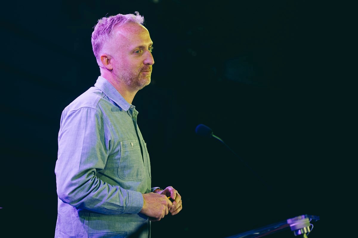 Tom Lawson standing on a stage under dramatic blue and purple lighting, wearing a light blue button‑up shirt. They are positioned near a lectern and microphone stand, holding their hands together as if mid‑talk. The dark background and spotlighting suggest a live presentation or speech at a professional event or conference.