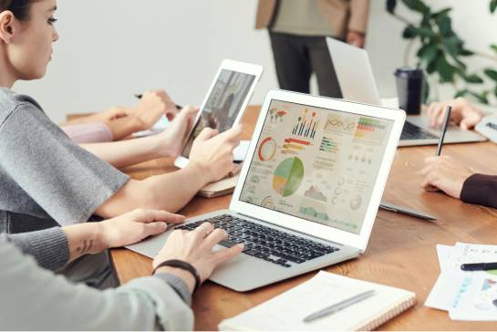 Four people in a meeting room work on laptops and tablets around a wooden table, displaying colorful charts and graphs, with a plant visible in the background.