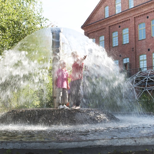 Two children standing under a dome-shaped water fountain near a brick building with large windows on a sunny day.