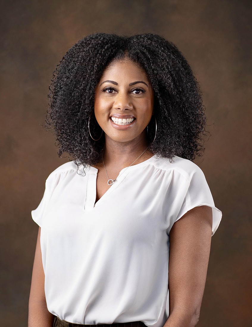 Smiling young woman with long hair standing against a dark green background, holding a finger to her chin.