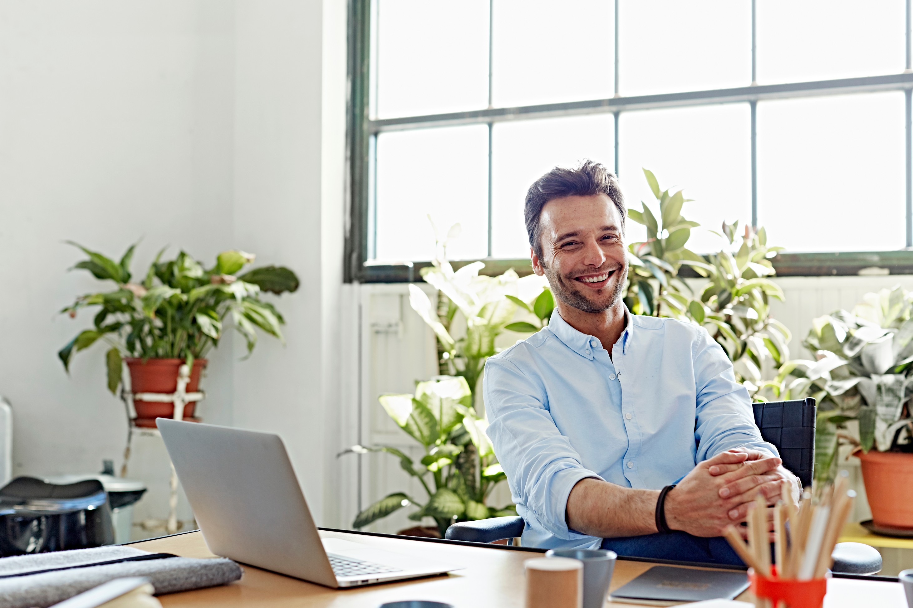 A retirement plan advisor smiles at a desk with an open laptop on the Pontera platform, notebook, coffee mug, and potted plants by large windows