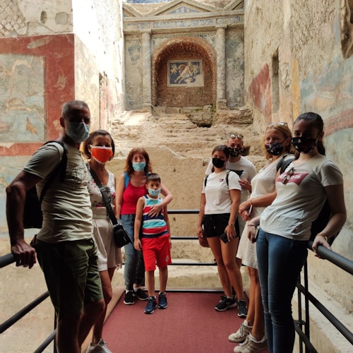 A group of people wearing masks stands on a ramp in front of ancient ruins with painted walls and an archway.