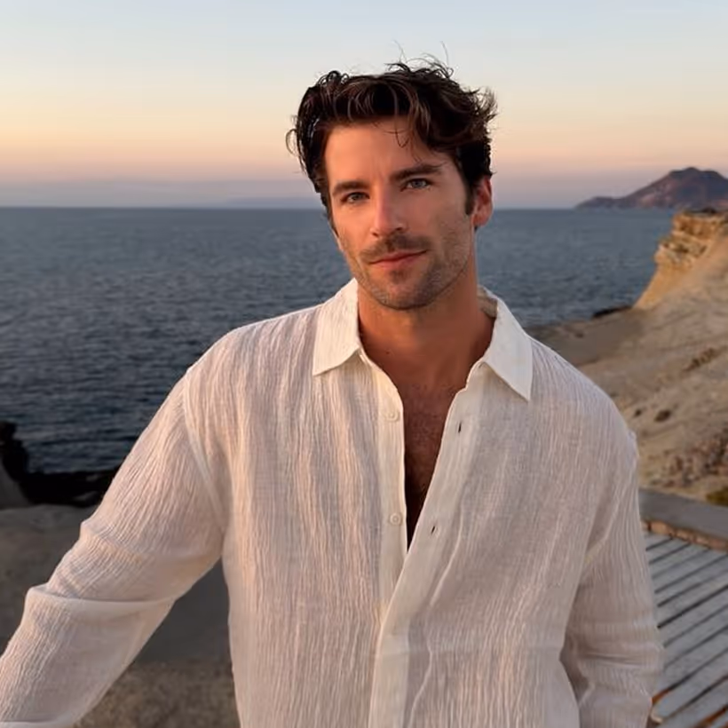 Man with tousled dark hair wearing an open white linen shirt by the ocean at sunset.