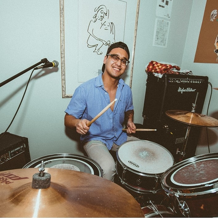 Ellixs playing drums while smiling at a camera