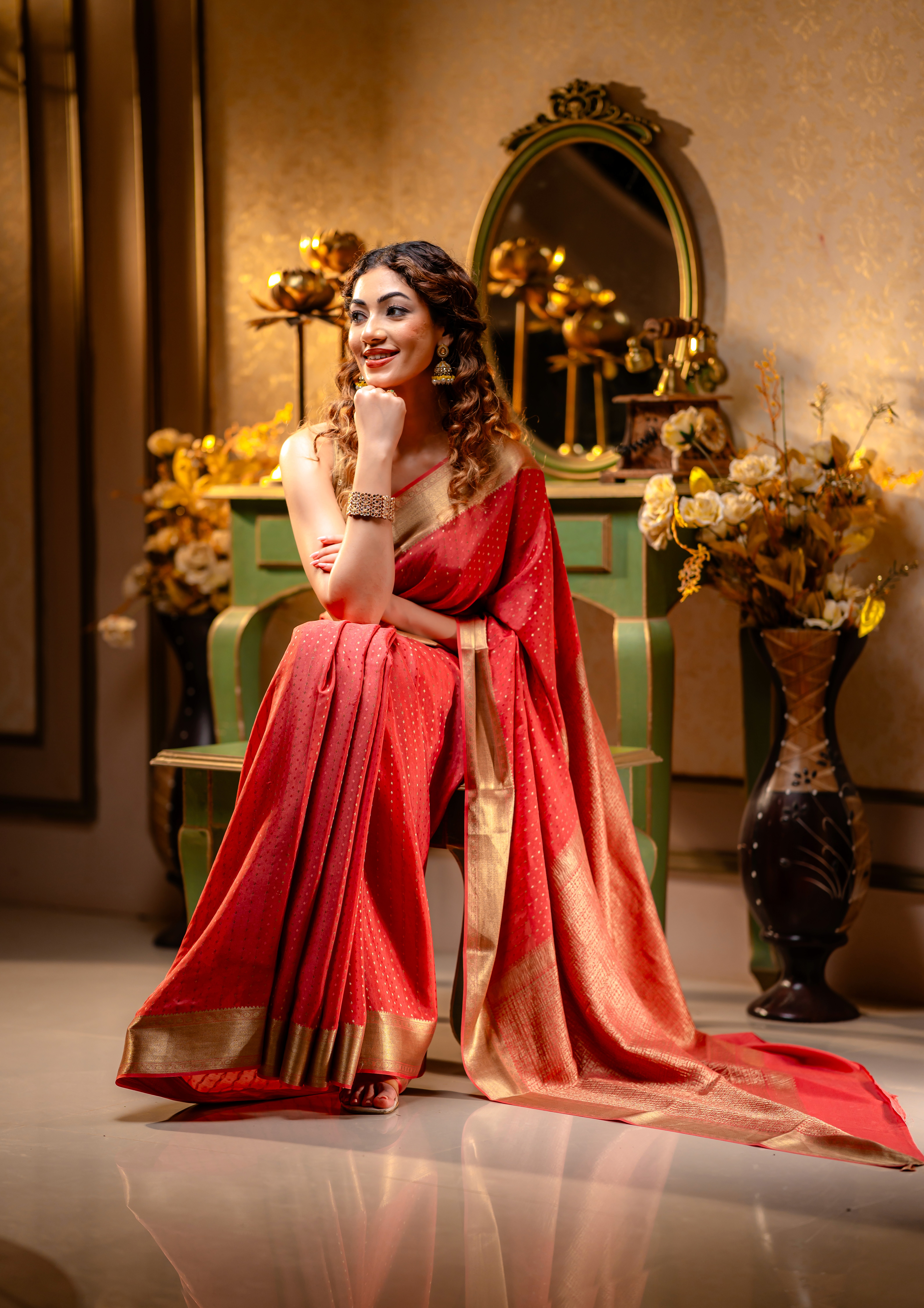 A woman wearing a red saree with golden borders from That Saree Shop, seated gracefully on a chair in an elegant room, highlighting the saree’s luxurious weave and classic festive charm.