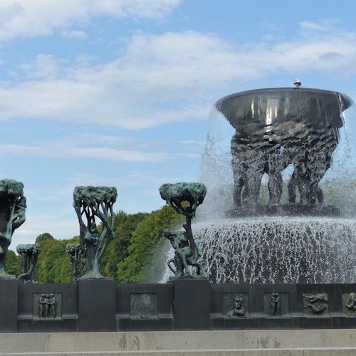 Бронзовые скульптуры фигур и деревьев окружают большой водный фонтан в парке, на фоне голубого неба с облаками.