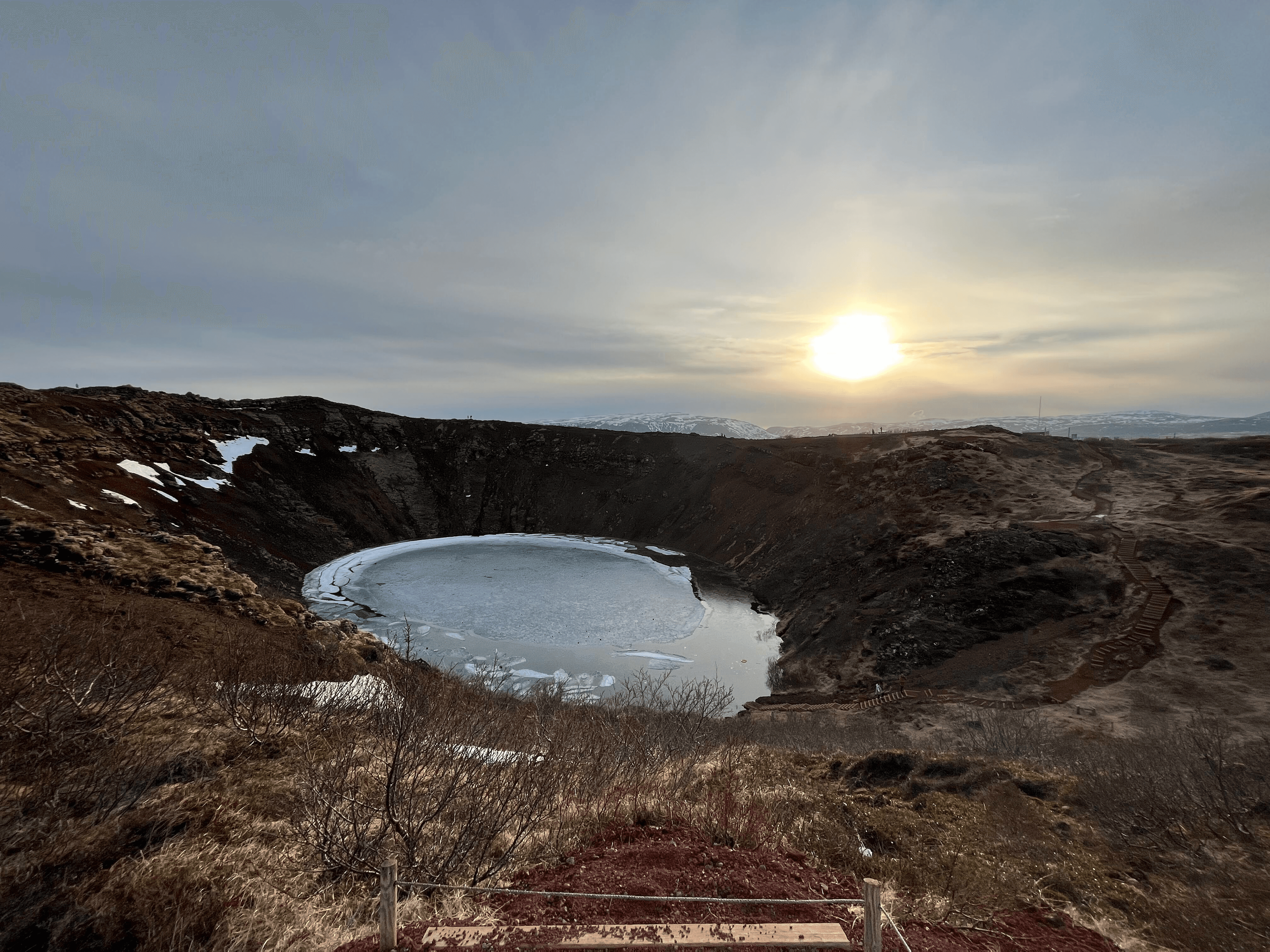 A frozen crater lake under a setting sun, surrounded by rugged terrain.