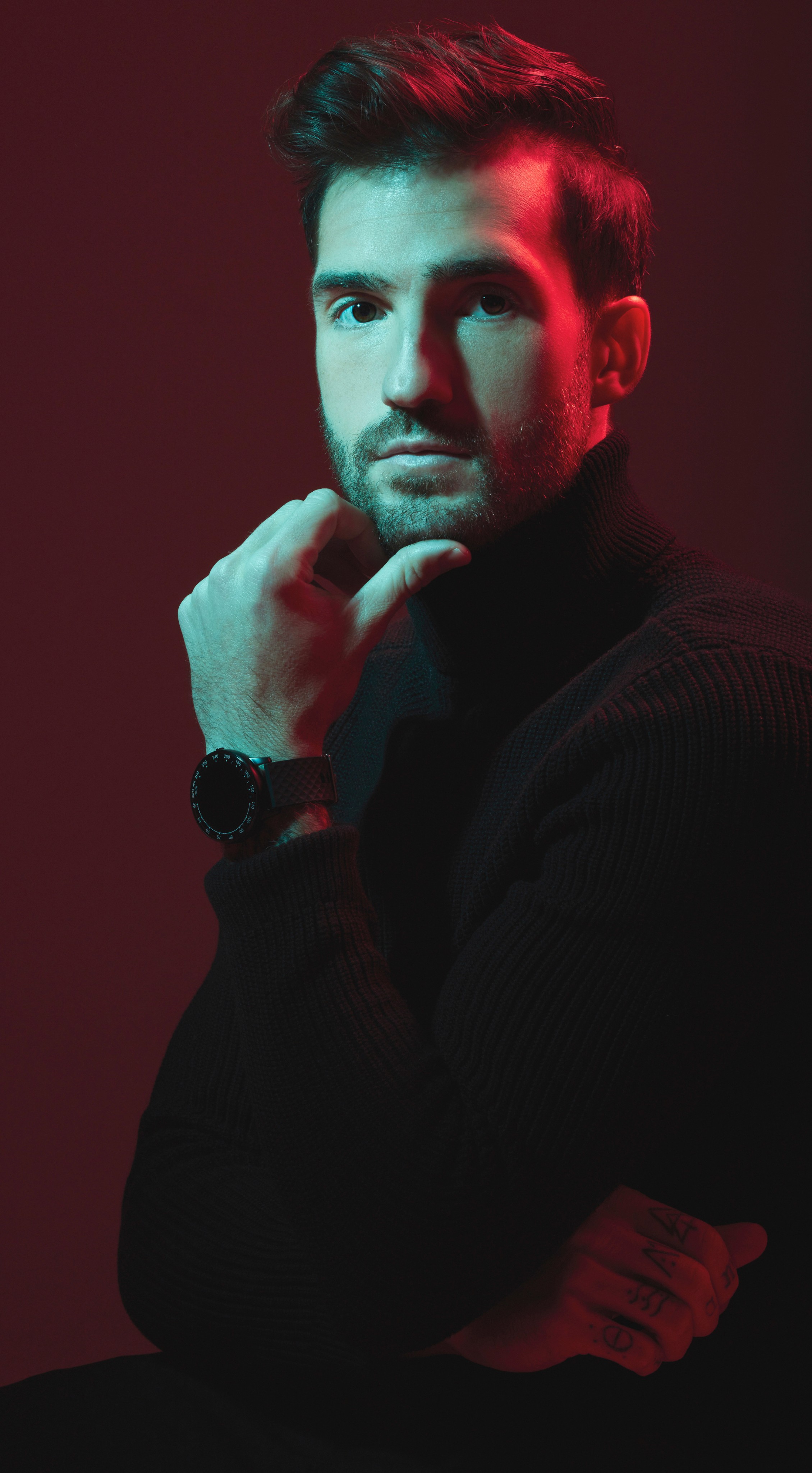 A cinematic studio portrait of a bearded man wearing a black turtleneck and a smartwatch. The photo features dramatic dual-tone lighting with teal highlights on one side and red ambient light on the other, creating a high-end, sophisticated editorial look.