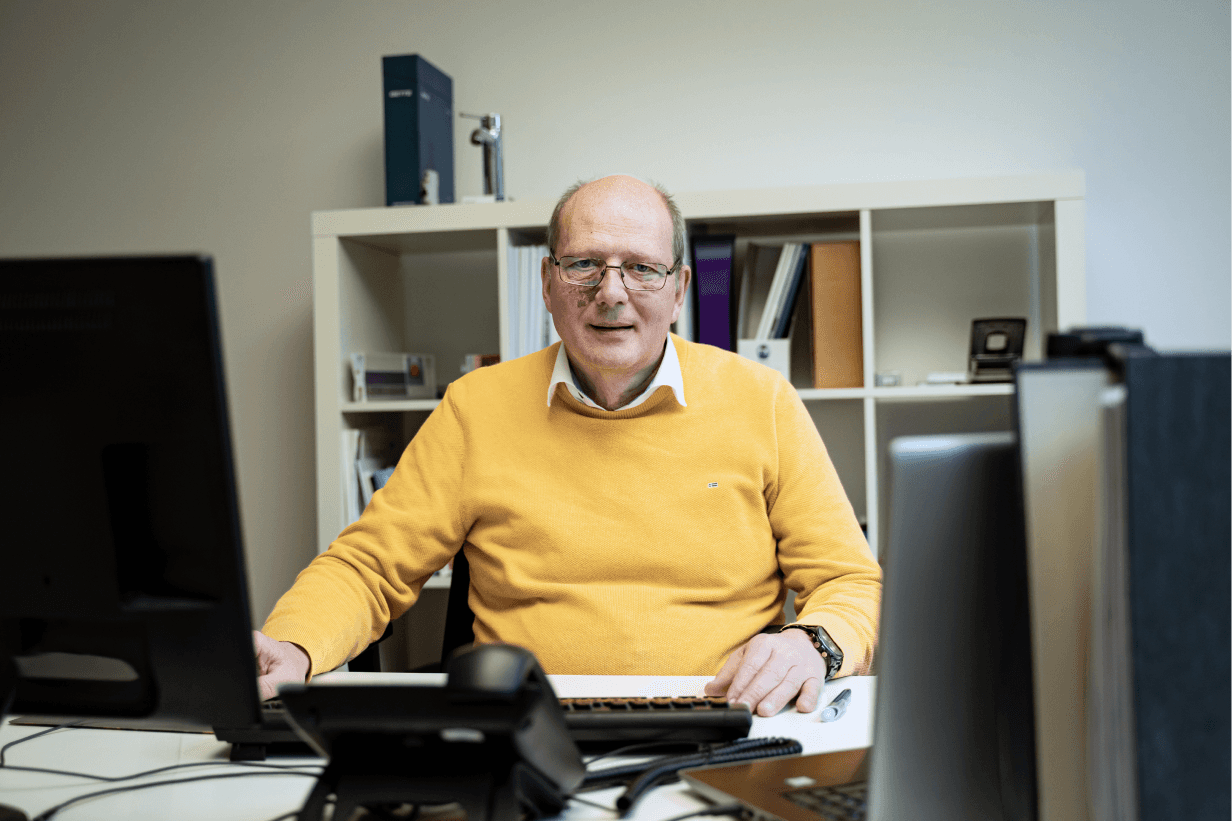 Älterer Mann im gelben Pullover sitzt am Schreibtisch im Büro der kba Architekten und Ingenieure GmbH, arbeitet am Computer und hat Regale mit Unterlagen im Hintergrund.