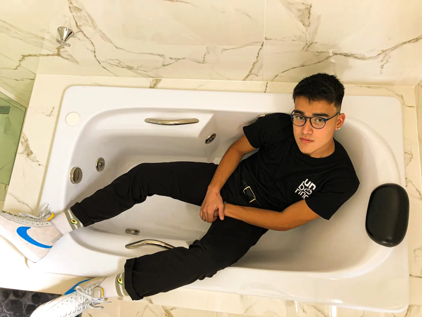 A top-down view of a young man in glasses reclining diagonally inside an empty white bathtub, wearing a black T-shirt with the stacked Unboring wordmark on the chest.