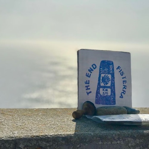 Un cartello che segna "La Fine Fisterra KM 0,0" si trova su una sporgenza, con l'acqua dell'oceano e la luce del sole visibili sullo sfondo.