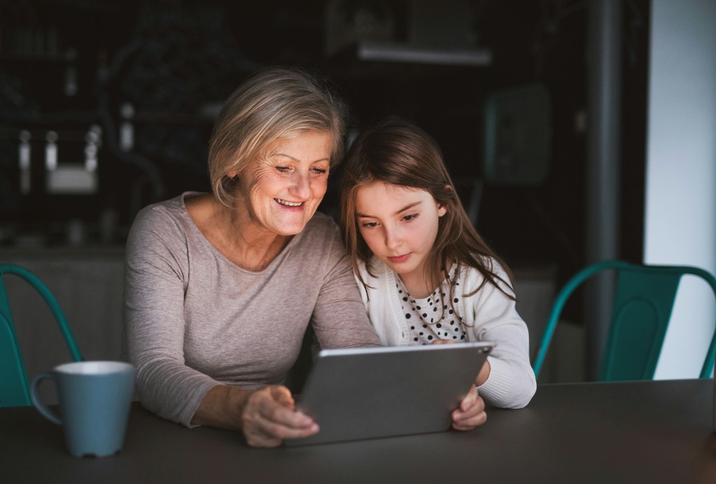 A smiling grandmother and her young granddaughter share a moment together while looking at a tablet, highlighting the joy of technology connecting different generations.