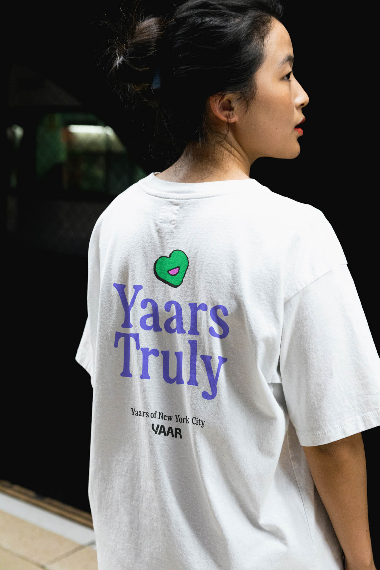 A person with dark hair pulled back in a bun is seen from the back, wearing a white t-shirt. The back of the shirt features the text "Yaars Truly" in blue, with a small green heart shape above the text. Below this, in smaller text, it reads "Years of New York City" and "VAAR". The background is dark and out of focus.