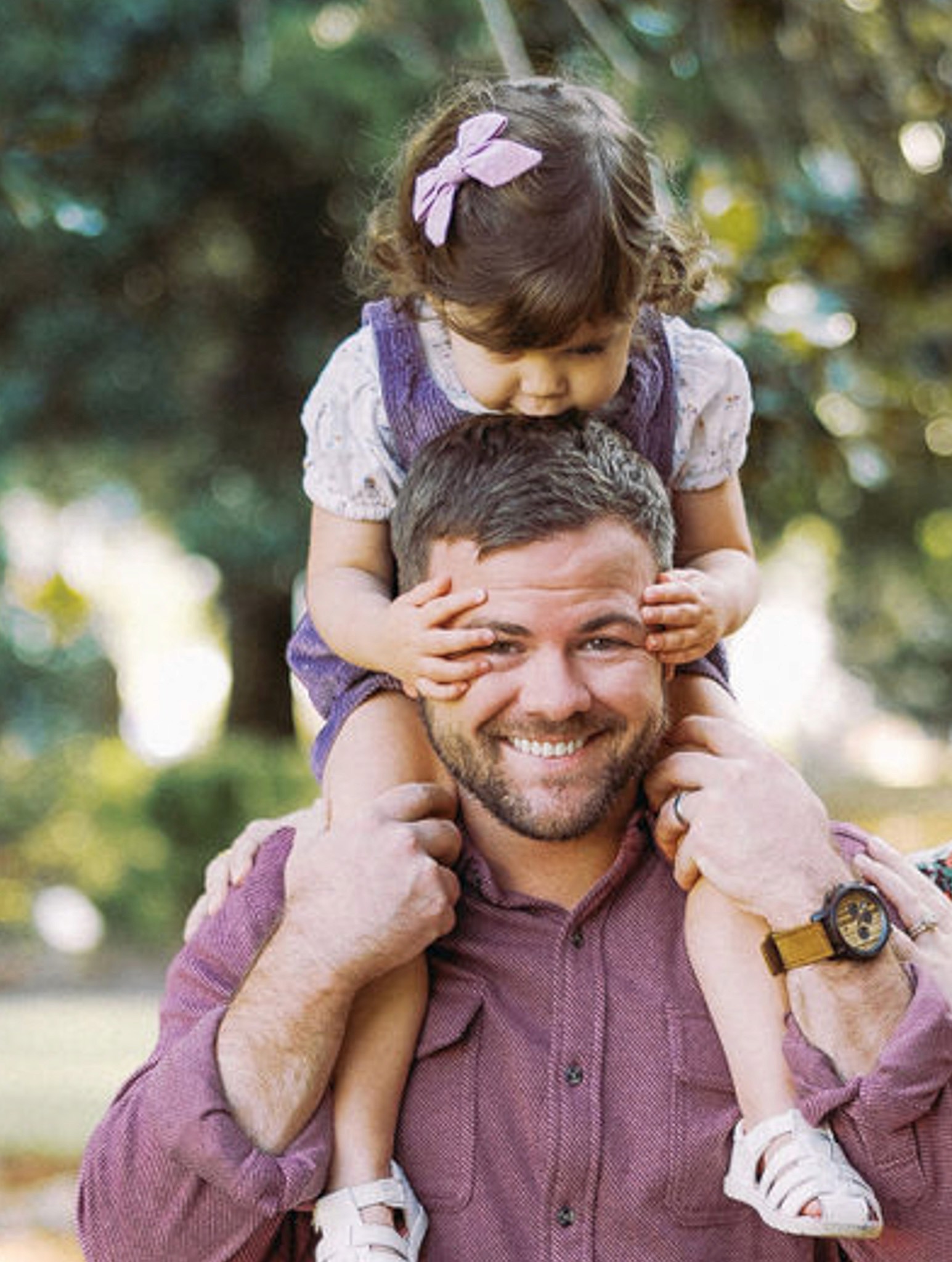A man smiles while carrying a girl on his shoulders, enjoying a sunny day outdoors. Green trees are in the background.