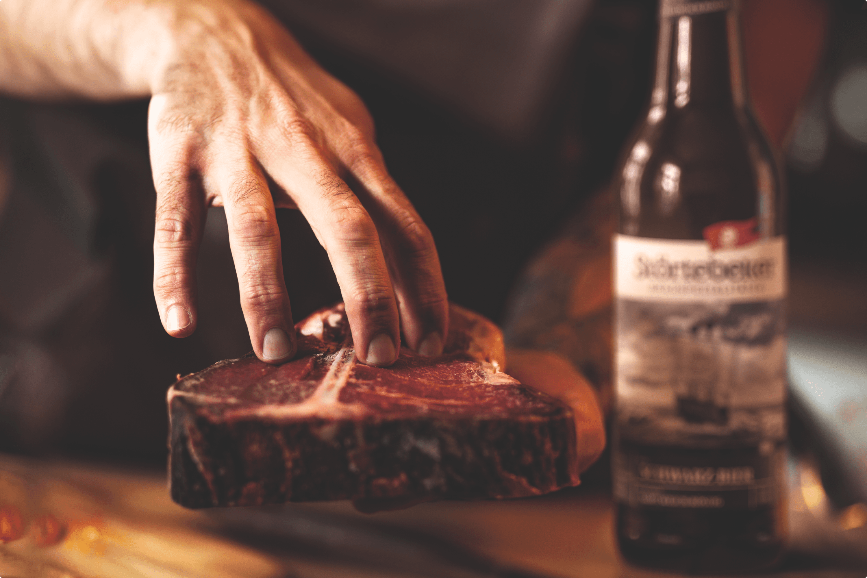 Hand touching t-bone steak next to beer bottle.