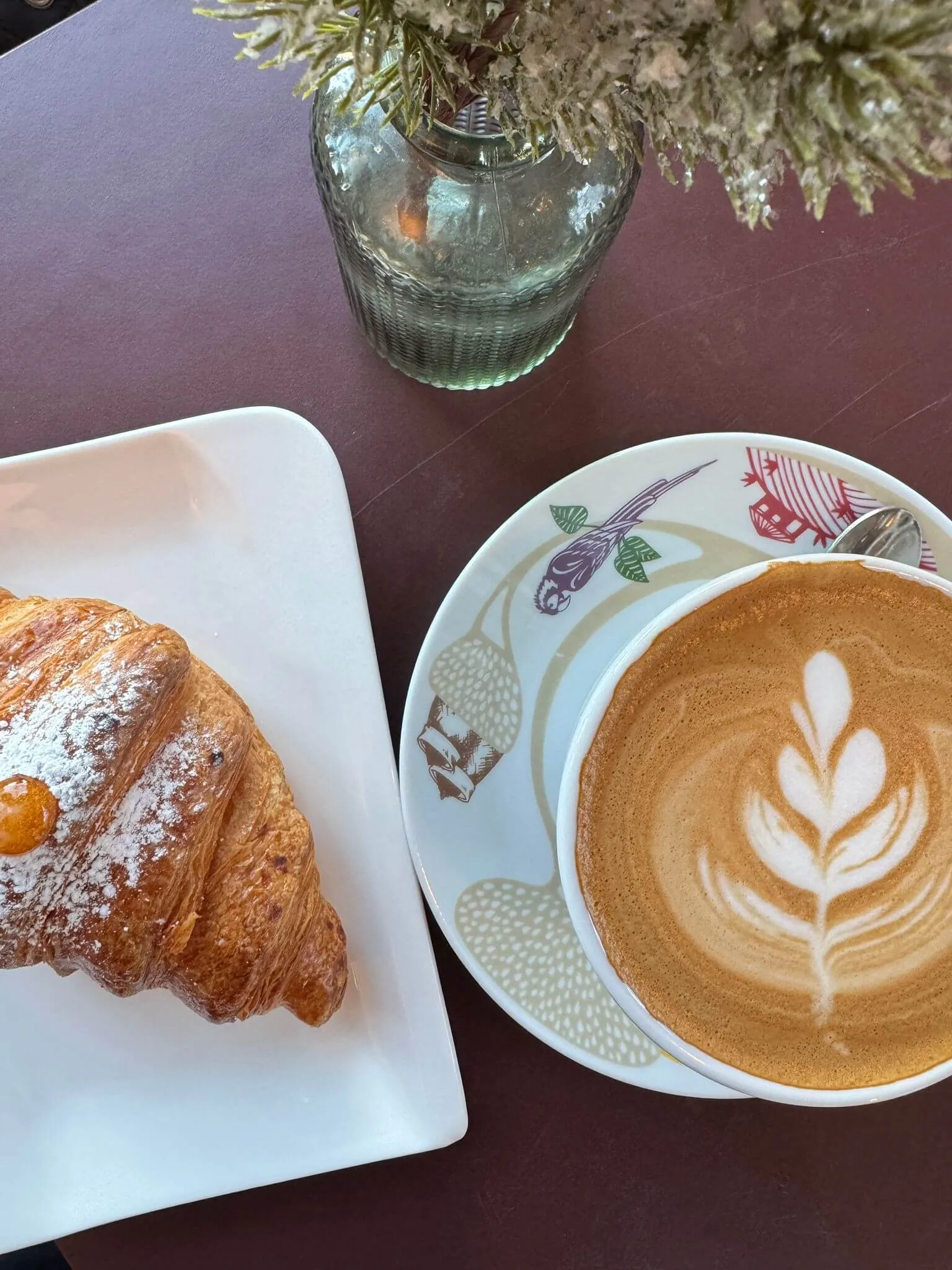 A croissant on a white plate beside a latte with a leaf design, set on a wooden table with flowers in the background.