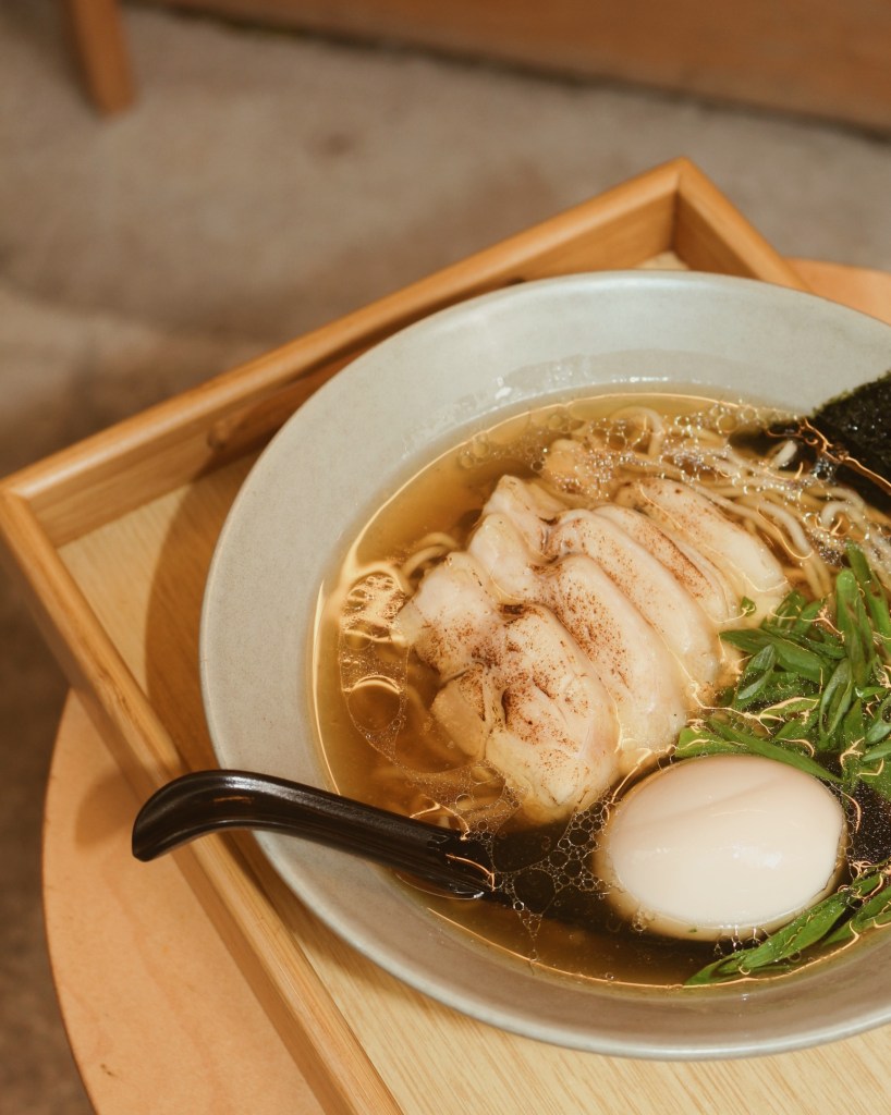 A bowl of clear chicken ramen featuring marinated chicken chashu, soft boiled egg, and green vegetables, served on a wooden tray.