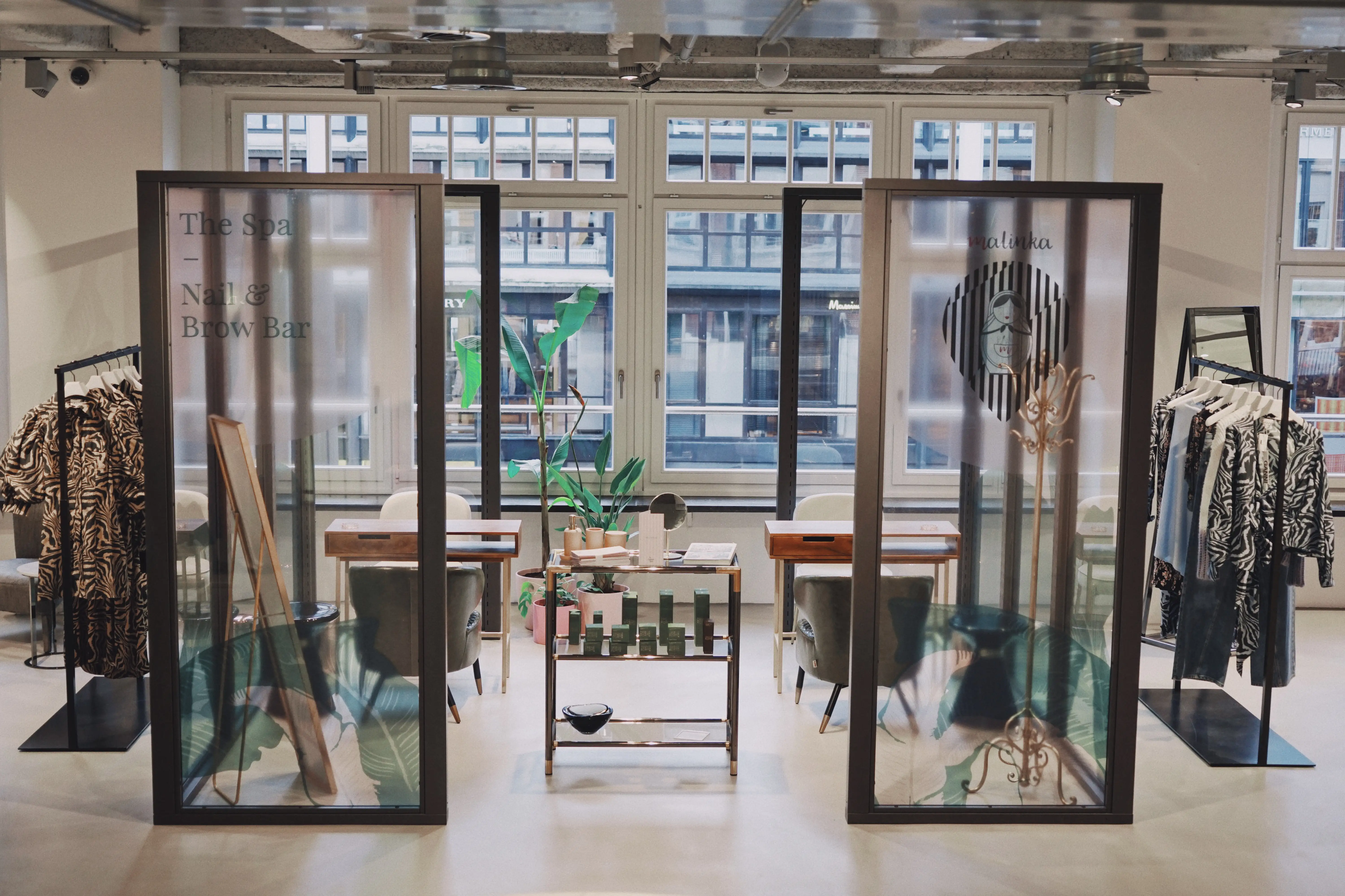 Spa and nail bar zone at UZWEI FASHION HOTEL framed by two glass partitions with lettering, manicure tables with chairs and large factory windows behind.