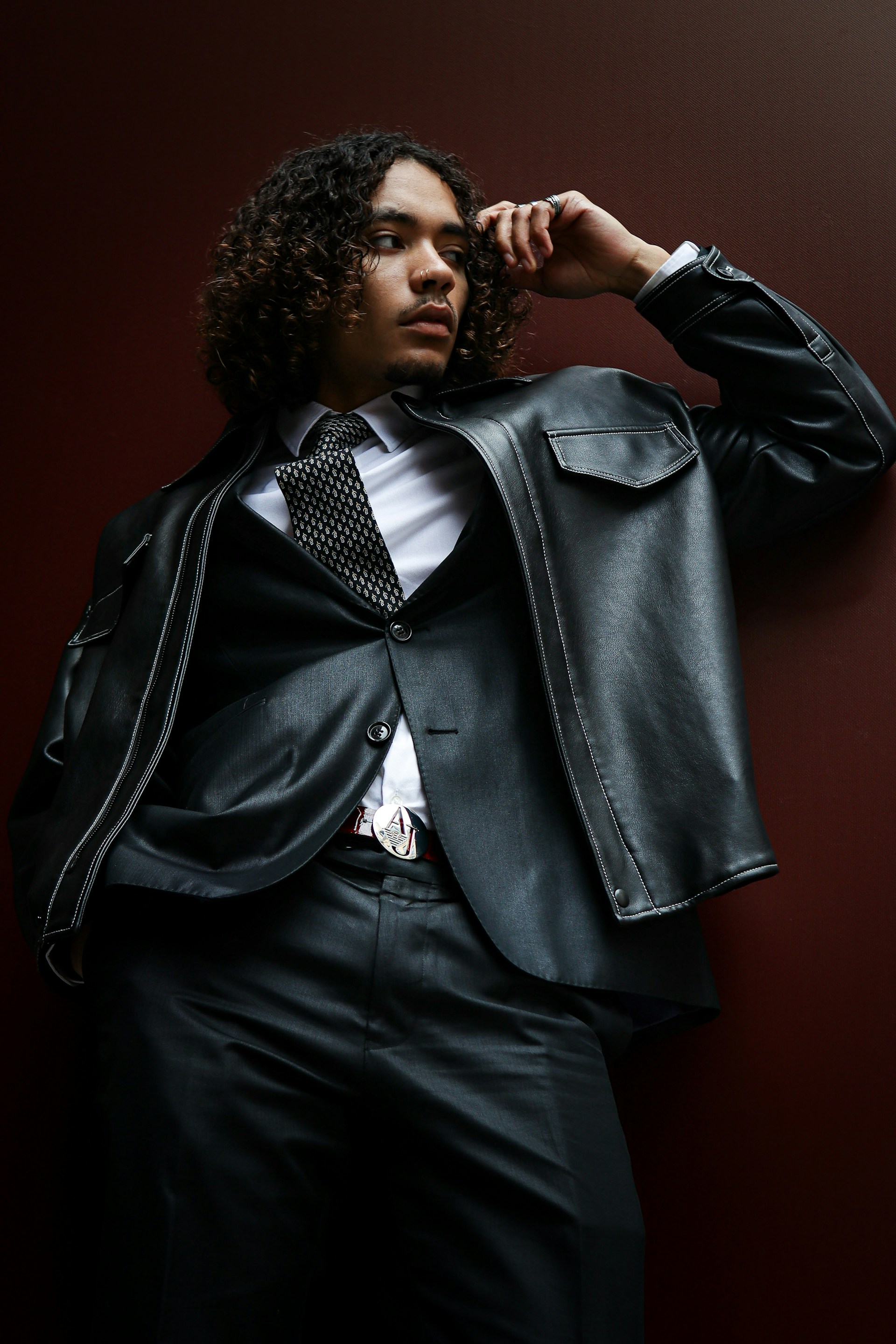 A person with long, curly brown hair leaning against a dark red wall in a low-angle shot. They are wearing a white dress shirt, a patterned black tie, a black blazer, and a black leather jacket with white stitching.