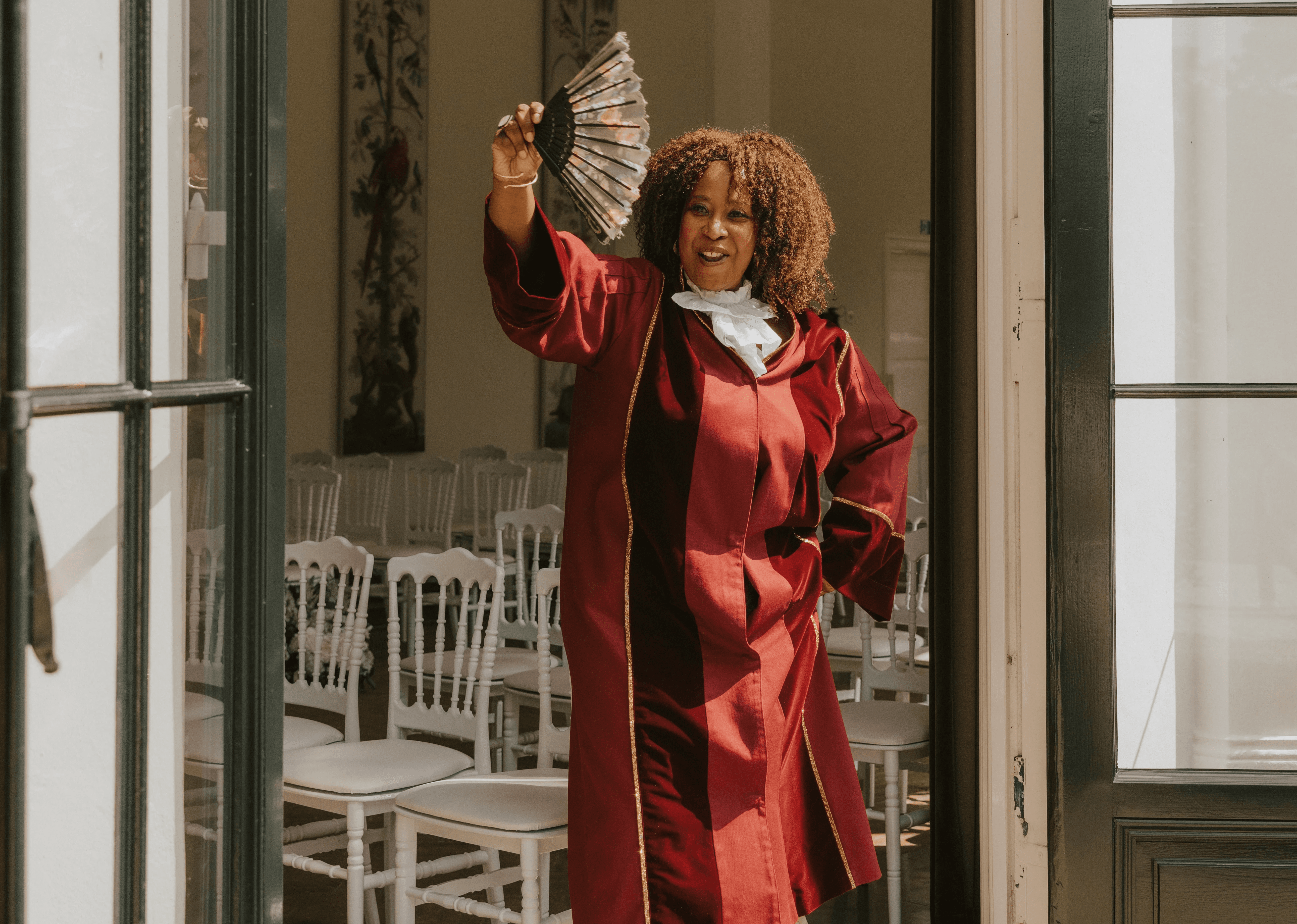 Een vrolijke zelfstandig trouwambtenaar in een bordeauxrode toga zwaait met een waaier tijdens een huwelijksceremonie, klaar om het bruidspaar te verwelkomen op hun trouwdag.