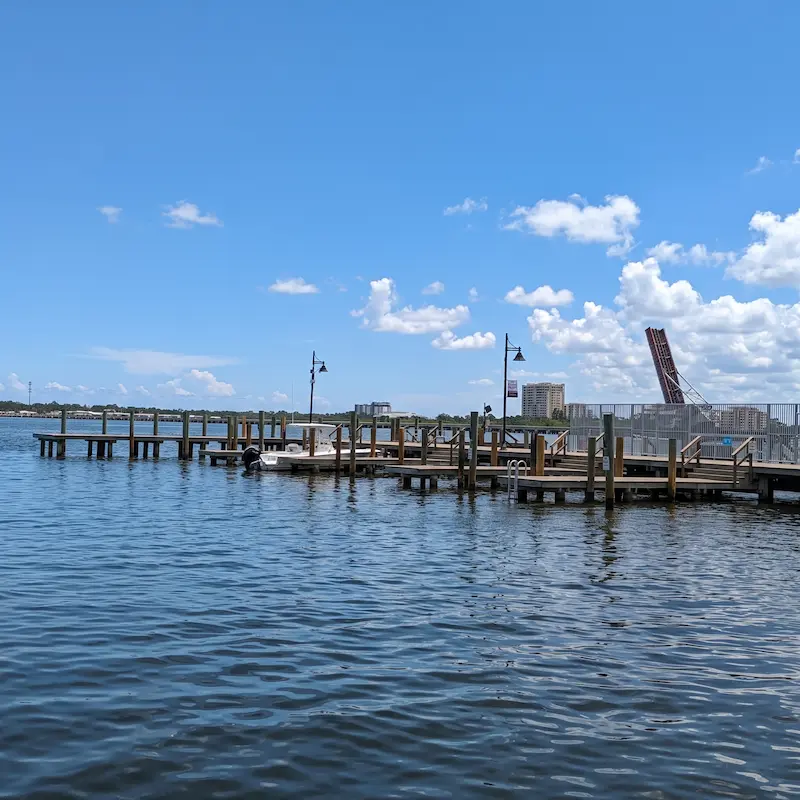 A vibrant playground at Riverwalk in Bradenton, FL features a variety of colorful play equipment including slides and swings, set amidst lush green artificial turf and shaded by large oak trees.