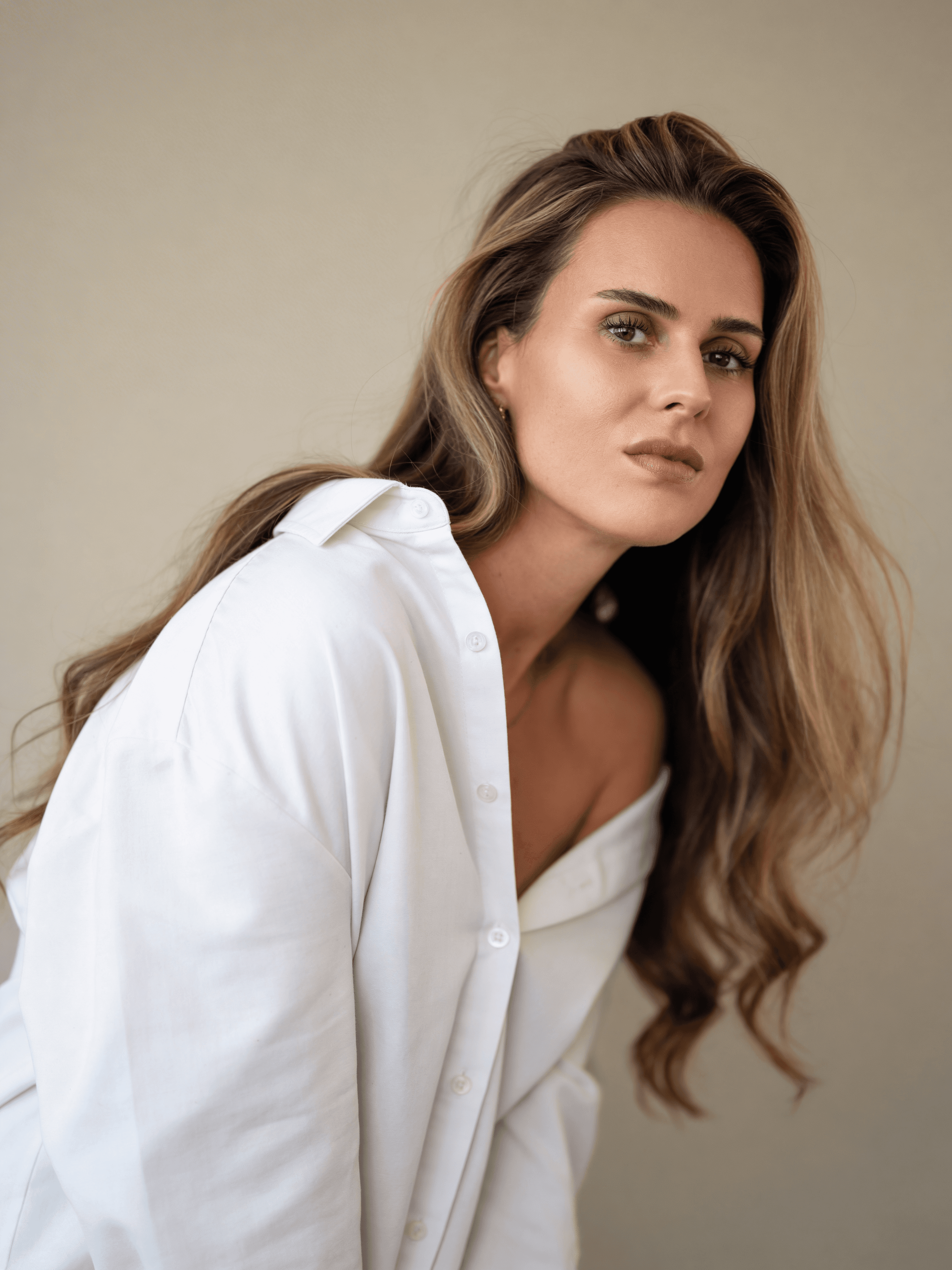 Weibliches KI-Model – Digitaler Zwilling von Trickhouse, weißes Oversize-Hemd, langes braun-blondes Haar, Blick über die Schulter, beiger Studio-Hintergrund, KI-Model-Agentur Düsseldorf.