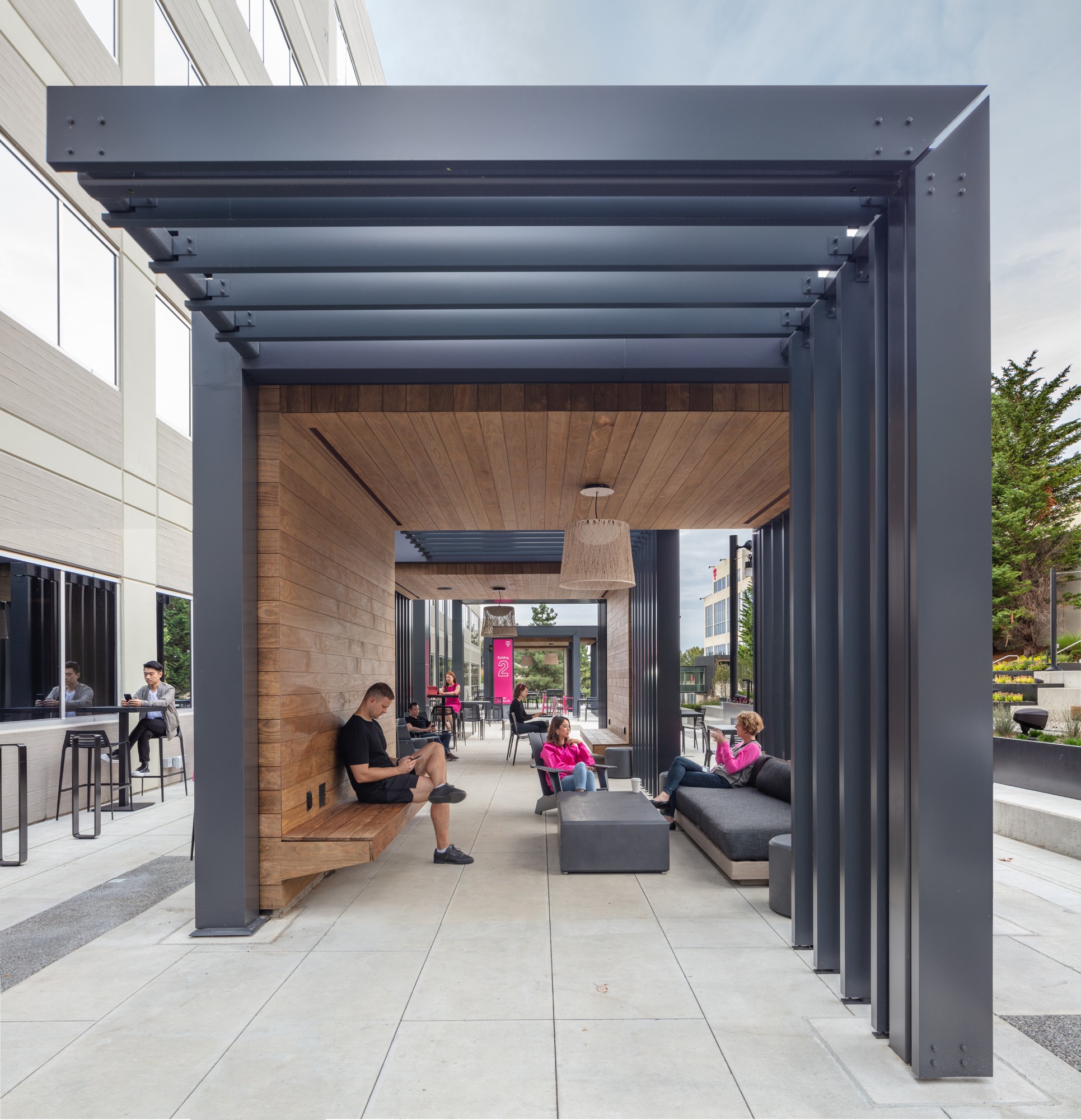 A side view of the outdoor "digit" pavilions, framed by L-shaped metal trellises that transition into solid wooden-clad enclosures, forming cozy lounges furnished with sofas, lighting, and ottomans.