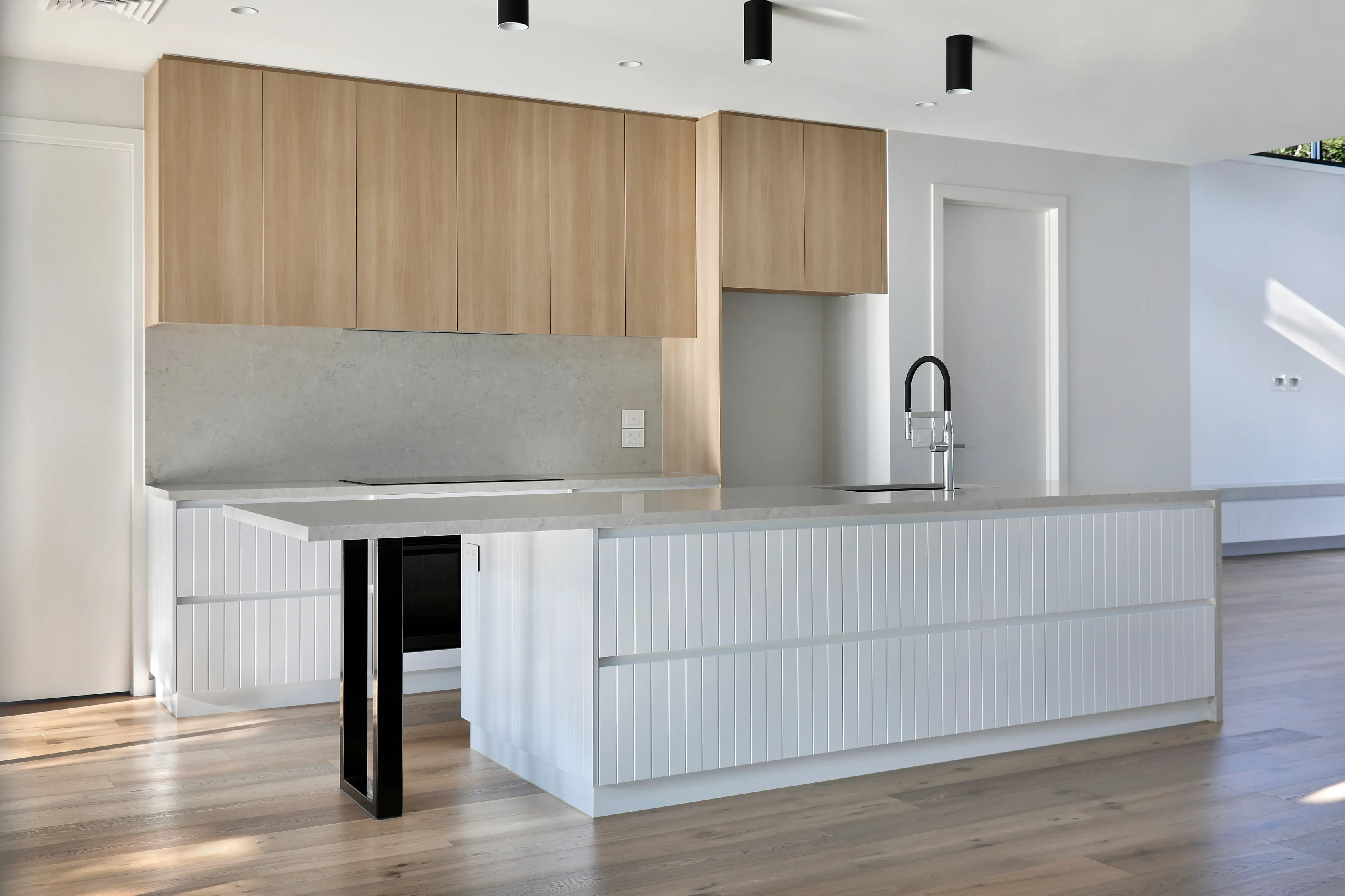 Kitchen with pale-oak cabinetry, a fluted white island with grey stone top on a black steel leg, black tap, integrated oven and induction cooktop over timber floors